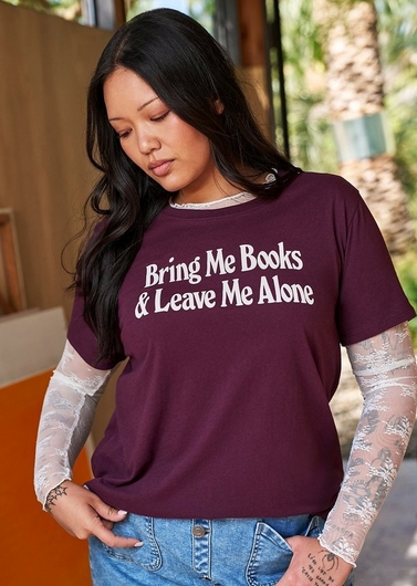 Photo of a woman with long straight black hair looking downward, standing in front of a blurred background with wood paneling, an orange wall section, and greenery with a palm tree. She wears a dark maroon graphic t-shirt with white text reading 'Bring Me Books & Leave Me Alone' layered over a white lace long sleeve top, paired with denim shorts. She has a visible tattoo with small text on her forearm.