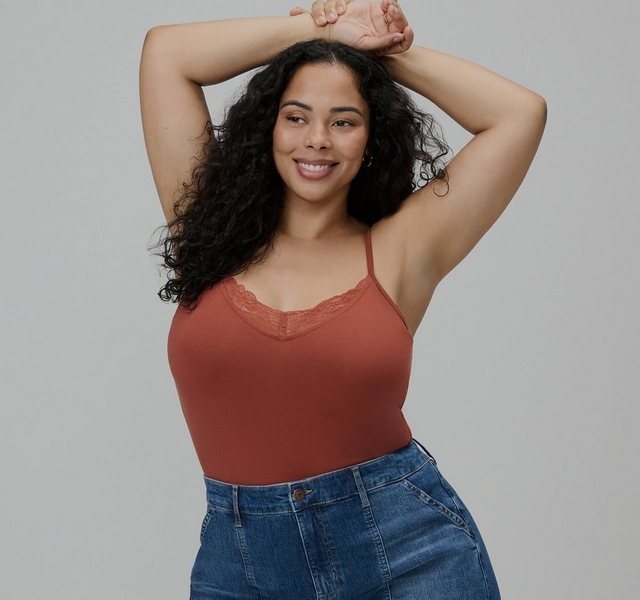 Photo of a woman with long dark curly hair against a plain light gray background, smiling with her head tilted and both arms raised above her head, wrists crossed together. She wears a rust orange cami top with a lace trimmed v-neckline and thin adjustable straps, paired with denim jeans visible at the waist. She has small hoop earrings.