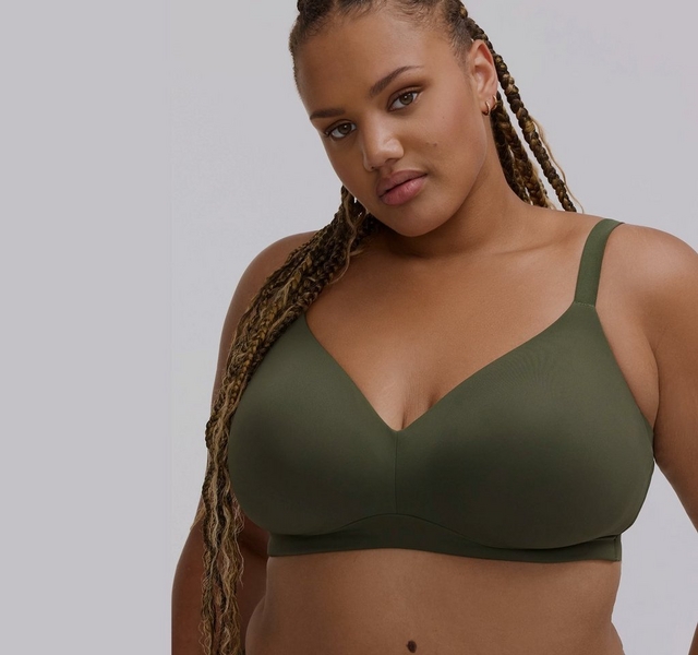Close-up photo of a woman with long braided hair in shades of brown and blonde against a plain light gray background. She wears an olive green wireless bra with a plunging v-neckline and thin straps. She has a small gold hoop earring visible and a beauty mark near her mouth.