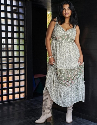  Photo of a woman with long dark wavy hair standing in front of a dark wooden slatted wall with a lattice patterned window structure to the left showing greenery outside. She wears a sleeveless maxi dress in a sage green and white mixed print combining small floral patterns with a paisley print panel at the waist and hem, featuring a sweetheart neckline, ruffled straps, and a tiered flowing skirt with ruffled trim. She has bracelets and rings on her hands and wears cream colored perforated pointed toe ankle boots with a stacked heel. One hand gathers up part of the skirt fabric.