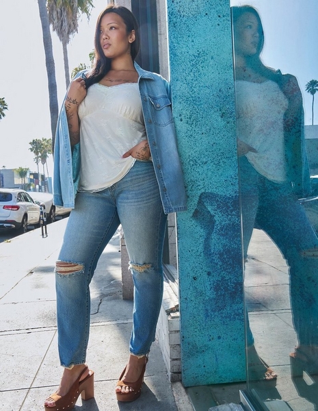 Photo of a woman with long straight dark hair standing on a sidewalk next to a teal textured wall with a reflective glass panel showing her mirrored reflection, with palm trees and parked cars visible in the background. She wears an open light denim shirt jacket over a white lace textured cami top, paired with light wash distressed skinny jeans with rips at the knees, and brown platform heeled sandals with ankle straps. She has visible tattoos on her arm and hand.