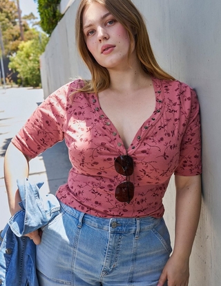 Close-up photo of a woman with light brown hair leaning against a gray wall with trees visible in the background. She wears a mauve pink top with a dark floral vine print, featuring elbow length sleeves and a deep v-neckline with a row of snap button closures down the front, paired with light wash blue jeans. Sunglasses hang from the front of her top, and she holds a light denim jacket in one hand.