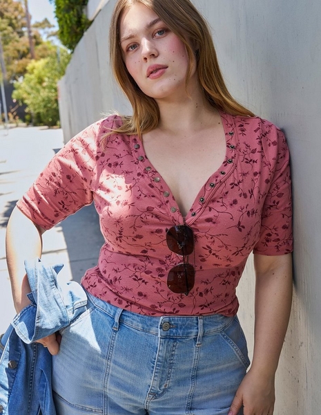 Close-up photo of a woman with light brown hair leaning against a gray wall with trees visible in the background. She wears a mauve pink top with a dark floral vine print, featuring elbow length sleeves and a deep v-neckline with a row of snap button closures down the front, paired with light wash blue jeans. Sunglasses hang from the front of her top, and she holds a light denim jacket in one hand.
