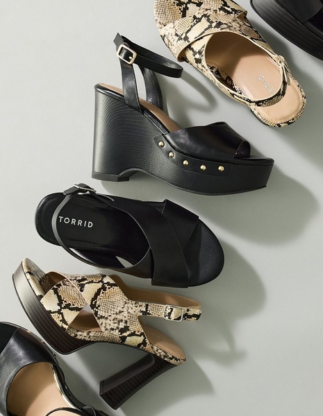 Close-up flat lay style photo of an assortment of platform sandals arranged on a light gray background. Included are a black leather wedge sandal with a crisscross front strap, an ankle strap, and gold stud details on the platform, a black leather sandal with a crisscross strap and a label reading 'Torrid' on the insole, and two snakeskin print sandals with crisscross straps and ankle buckle closures, one shown from above and one in the foreground with a stacked wooden heel.