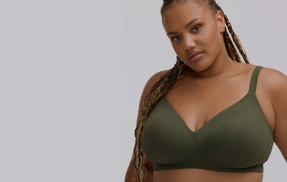 Photo of a woman with long braided hair in shades of brown and blonde against a plain light gray background. She wears an olive green wireless bra with a plunging v-neckline and thin straps. She has small gold hoop earrings and a beauty mark near her mouth.