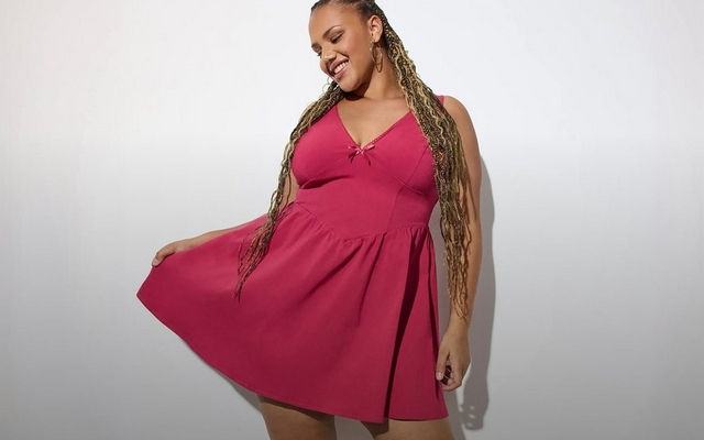 A model with long dark hair and visible arm tattoos poses against a light grey background wearing a yellow floral ruffle camisole top with spaghetti straps and layered gold necklaces, one arm raised behind her head.