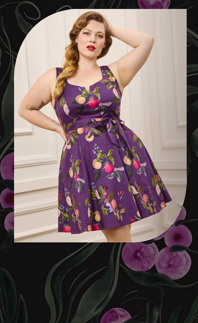 Close-up vertical photo of a woman with long wavy strawberry blonde hair styled in a vintage pin curl look, set within an arched frame against a dark illustrated background of green leaves and purple calla lily flowers. She poses with one hand on her hip and the other touching her hair, wearing a purple fit and flare dress with a sweetheart neckline, a v-shaped keyhole cutout, and an allover fruit print featuring peaches, plums, cherries, and berries with green leaves, cinched with a matching fabric tie belt. She has a visible tattoo on her forearm and wears a ring.