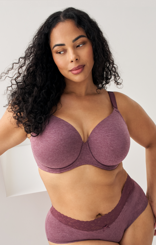 Close-up photo of a woman with long dark curly hair against a plain light gray background, looking off to the side with one hand resting on her hip. She wears a heathered mauve underwire bra with adjustable straps and matching lace trimmed high waisted underwear, with a visible tattoo near her hip.