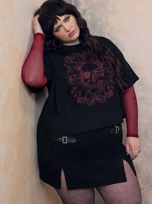 Close-up photo of a woman with dark wavy hair and bangs posing against a mottled beige and terracotta textured wall, one hand resting in her hair. She wears red fishnet sleeves layered under a black cropped t-shirt featuring a red skull graphic surrounded by roses and spiderweb detailing, paired with a black mini skirt with multiple black buckle straps with gold hardware and a front slit, and fishnet tights, along with hoop earrings and rings on her fingers.