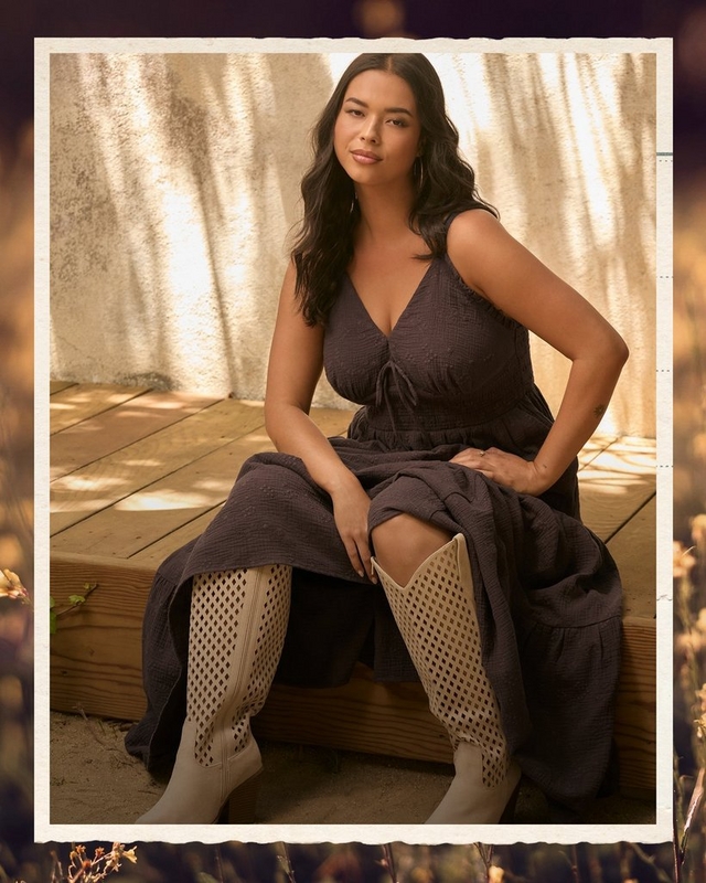 Photo of a woman with long dark wavy hair sitting on a wooden deck bench in dappled sunlight, framed with a white deckled border. She wears a dark brown textured sleeveless midi dress with a tie front v-neckline and an empire waist, paired with cream perforated knee-high zip boots, one hand on her hip and the other resting on her raised knee, with a visible tattoo near her wrist.