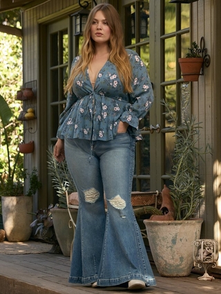 Photo of a woman with long wavy strawberry blonde hair standing on a porch with potted plants and French doors in the background. She wears a dusty blue floral print blouse with a plunging tie front neckline, bishop sleeves with ruffled cuffs, and a peplum hem, paired with light wash distressed flare jeans.