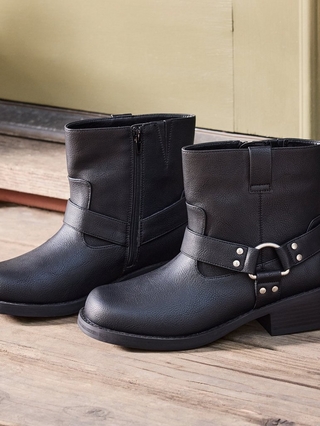 Close-up photo of a pair of black faux leather ankle boots with a stacked block heel, one boot showing a side zipper and the other featuring a decorative harness strap with a silver ring and stud detailing, placed on a wooden porch floor near a green painted door frame.