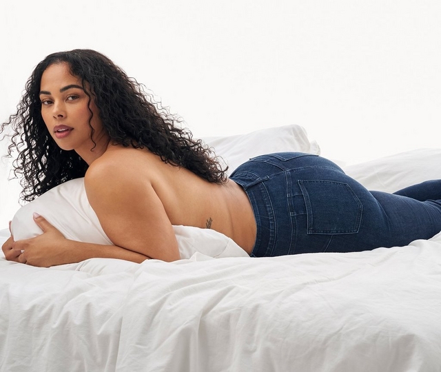 A woman with long curly dark brown hair lies on her side on a bed with white bedding, facing slightly toward the camera. She wears dark indigo skinny jeans and is topless, holding a white pillow against her chest with both arms to cover herself. Her legs are bent and crossed at the ankles. The background is plain white, keeping the focus on the jeans she is wearing.