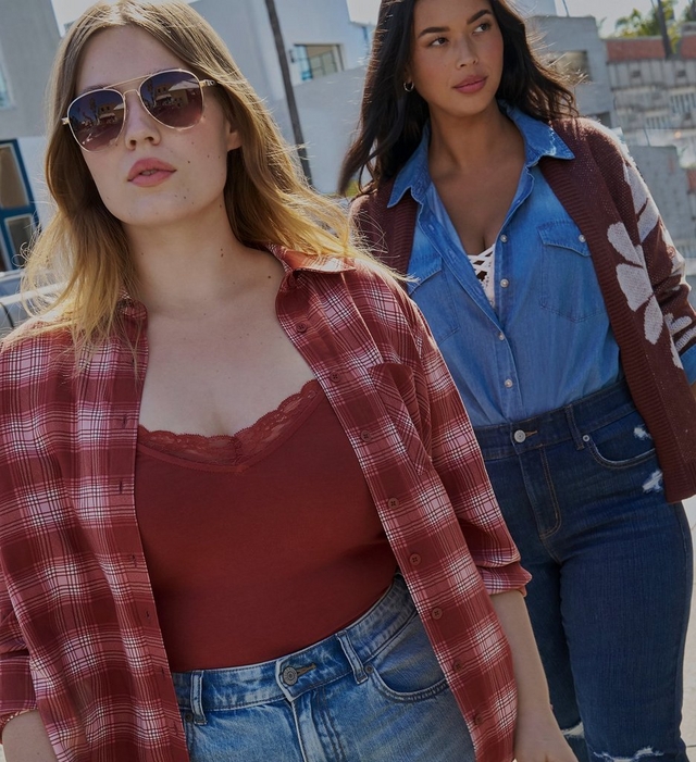 Two women walk outdoors on a city street with blurred buildings and greenery in the background. In the foreground, a woman with long wavy blonde hair wears sunglasses, a rust red and white plaid flannel shirt worn open over a burgundy lace trimmed cami, and denim jeans. Behind her, a woman with dark hair wears a chambray denim button up shirt layered under a brown cardigan with a large white floral design on the sleeve, paired with dark denim jeans.