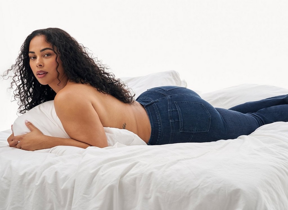 A woman with long curly dark brown hair lies on her side on a bed with white bedding, facing slightly toward the camera. She wears dark indigo skinny jeans and is topless, holding a white pillow against her chest with both arms to cover herself. Her legs are bent and crossed at the ankles. The background is plain white, keeping the focus on the jeans she is wearing.