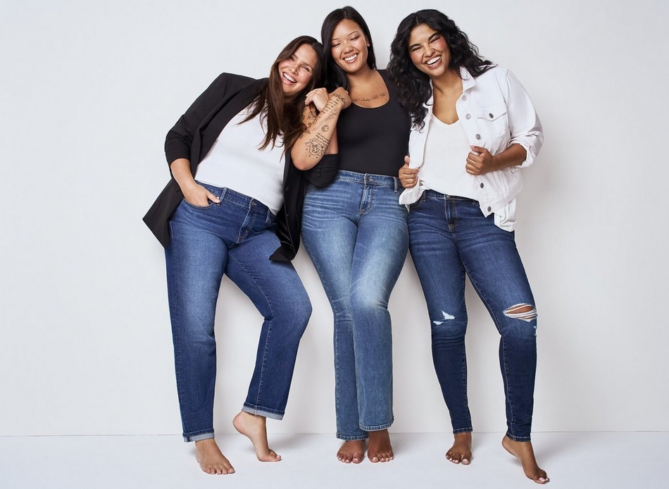 Three women stand together against a white background, all wearing dark wash denim jeans and posing barefoot. The woman on the left wears a black blazer over a white top with her hand in her pocket. The woman in the middle wears a black tank top and has her hands raised playfully. The woman on the right wears a white button up shirt with distressed ripped jeans.