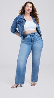 Photo of a woman with long wavy brown hair against a plain light gray background, wearing a white cropped tube top under an open medium wash denim jacket, paired with light wash straight leg jeans and silver strappy heeled sandals. 