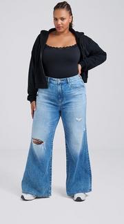 Photo of a woman with dark hair pulled back against a plain light gray background, wearing a black lace trimmed cami top under an open black zip-up hoodie, paired with medium wash distressed wide leg jeans with a ripped knee and white sneakers, one hand on her hip. 