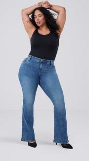 Photo of a woman with long curly dark hair against a plain light gray background, both arms raised above her head with wrists crossed, wearing a black sleeveless top paired with medium wash bootcut jeans and black pointed toe heels. 