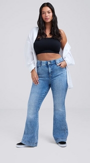 Photo of a woman with long dark wavy hair against a plain light gray background, wearing a black cropped tube top under an open white button-up shirt, paired with medium wash flare jeans and black platform sneakers, one hand in her pocket. 