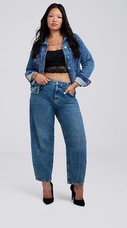 Photo of a woman with long dark hair against a plain light gray background, wearing a black lace trimmed cami top under an open medium wash denim jacket, paired with medium wash barrel leg jeans and black pointed toe heels, with visible neck and hand tattoos. 