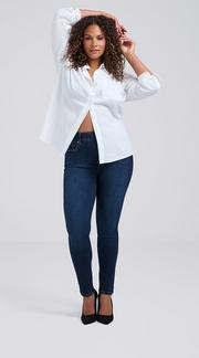 Photo of a woman with long curly dark hair against a plain light gray background, both arms raised with hands behind her head, wearing an open white button-up shirt paired with dark wash skinny jeans and black pointed toe heels.
