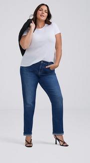 Photo of a woman with long dark wavy hair against a plain light gray background, holding a black jacket over one shoulder, wearing a white t-shirt paired with medium wash straight leg jeans with cuffed hems and black strappy heeled sandals. 