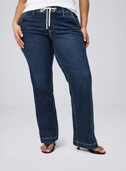 Cropped photo showing a woman's lower body against a plain light gray background, wearing a white top with dark wash wide leg jeans featuring a drawstring waistband, paired with black strappy heeled sandals. 