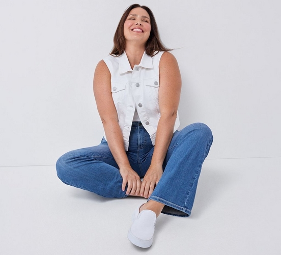 A woman with shoulder length brown hair sits cross legged on a white background, laughing with her eyes closed. She wears a light wash sleeveless denim jacket over a white top, medium wash straight leg jeans, and white sneakers. Her hands rest together in front of her. 