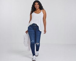 A woman with curly dark hair and light brown skin smiles while walking, wearing a white spaghetti strap tank top tucked into dark blue ripped skinny jeans with distressing at the knee. She carries a cream colored jacket over one arm and wears white sneakers with black and gray accents. The background is plain light gray.