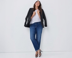 A woman with wavy brown hair and light skin leans against a wall with her head tilted back and eyes closed, smiling. She wears an open black blazer over a white t-shirt, paired with dark blue straight leg jeans cuffed at the ankle, and black strappy high heeled sandals. The background is plain light gray.