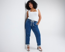 A woman with curly dark hair and light brown skin leans against a wall with one hand in her pocket, looking at the camera. She wears a white sleeveless top with a square neckline tucked into medium wash blue denim pants with a white drawstring tie at the waist and cuffed hems. She wears white platform block heel sandals. The background is plain light gray.