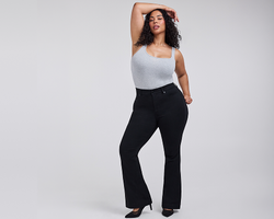 A woman with curly dark hair and medium skin tone poses with one arm raised above her head, resting on her head. She wears a light gray ribbed tank top with a square neckline tucked into black flared trousers, paired with black pointed toe high heels. The background is plain light gray.