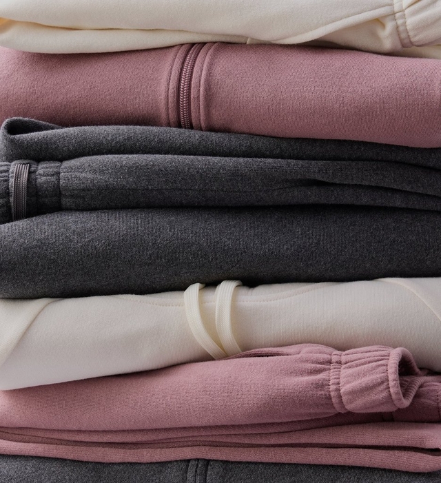 Close-up stack of folded fleece sweatshirts and joggers layered in cream, mauve pink, heather gray, and cream again, with mauve pink pieces visible at the bottom. Visible details include a ribbed cuff, a zipper pull, and a cream colored drawstring threaded through one garment. No people or text appear in the image.