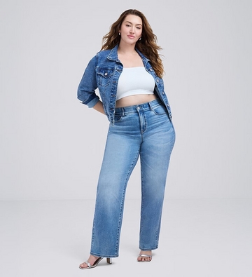Photo of a woman with long wavy brown hair against a plain light gray background, wearing a white cropped tube top under an open medium wash denim jacket, paired with light wash straight leg jeans and silver strappy heeled sandals. 