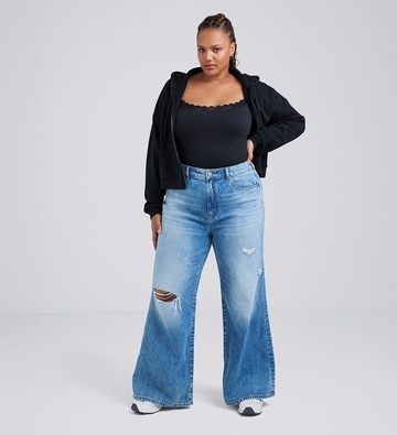 Photo of a woman with dark hair pulled back against a plain light gray background, wearing a black lace trimmed cami top under an open black zip-up hoodie, paired with medium wash distressed wide leg jeans with a ripped knee and white sneakers, one hand on her hip. 