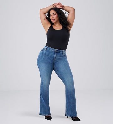 Photo of a woman with long curly dark hair against a plain light gray background, both arms raised above her head with wrists crossed, wearing a black sleeveless top paired with medium wash bootcut jeans and black pointed toe heels. 