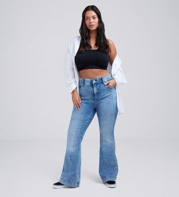 Photo of a woman with long dark wavy hair against a plain light gray background, wearing a black cropped tube top under an open white button-up shirt, paired with medium wash flare jeans and black platform sneakers, one hand in her pocket. 