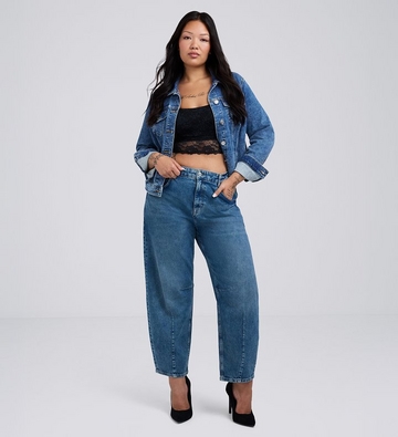 Photo of a woman with long dark hair against a plain light gray background, wearing a black lace trimmed cami top under an open medium wash denim jacket, paired with medium wash barrel leg jeans and black pointed toe heels, with visible neck and hand tattoos. 