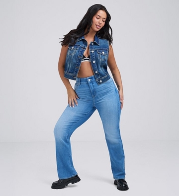Photo of a woman with long dark hair against a plain light gray background, mid stride with one hand on her hip, wearing a black and white striped bralette under an open sleeveless medium wash denim vest, paired with medium wash bootcut jeans and black chunky loafers. 