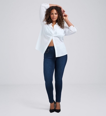 Photo of a woman with long curly dark hair against a plain light gray background, both arms raised with hands behind her head, wearing an open white button-up shirt paired with dark wash skinny jeans and black pointed toe heels.