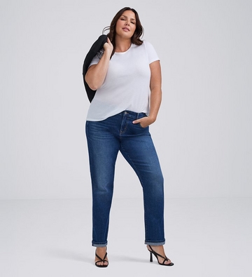 Photo of a woman with long dark wavy hair against a plain light gray background, holding a black jacket over one shoulder, wearing a white t-shirt paired with medium wash straight leg jeans with cuffed hems and black strappy heeled sandals. 