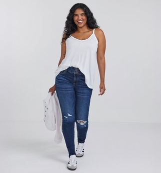 A woman with curly dark hair and light brown skin smiles while walking, wearing a white spaghetti strap tank top tucked into dark blue ripped skinny jeans with distressing at the knee. She carries a cream colored jacket over one arm and wears white sneakers with black and gray accents. The background is plain light gray.