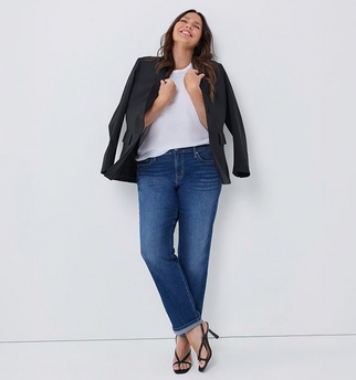 A woman with wavy brown hair and light skin leans against a wall with her head tilted back and eyes closed, smiling. She wears an open black blazer over a white t-shirt, paired with dark blue straight leg jeans cuffed at the ankle, and black strappy high heeled sandals. The background is plain light gray.