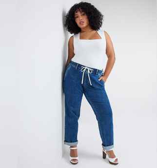 A woman with curly dark hair and light brown skin leans against a wall with one hand in her pocket, looking at the camera. She wears a white sleeveless top with a square neckline tucked into medium wash blue denim pants with a white drawstring tie at the waist and cuffed hems. She wears white platform block heel sandals. The background is plain light gray.