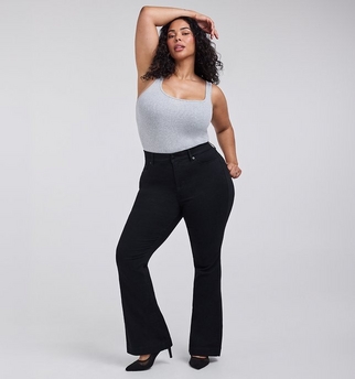 A woman with curly dark hair and medium skin tone poses with one arm raised above her head, resting on her head. She wears a light gray ribbed tank top with a square neckline tucked into black flared trousers, paired with black pointed toe high heels. The background is plain light gray.