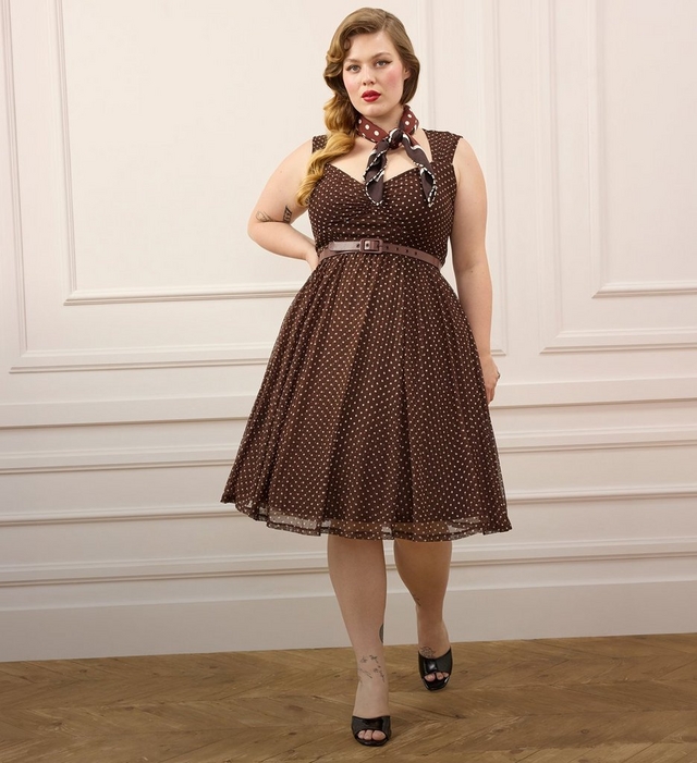 A woman with curled honey blonde hair styled in vintage waves stands with one hand on her hip, wearing a brown polka dot fit and flare dress with a sweetheart neckline, thin straps, and a brown belt at the waist. She has a polka dot scarf tied around her neck, red lipstick, and a small tattoo on her forearm. She wears black open toe heeled sandals and has a tattoo visible on her ankle. She stands on a herringbone wood floor in front of a white paneled wall.
