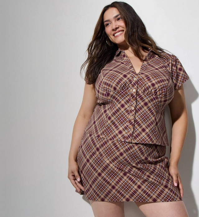 Close-up view of a smiling woman with long wavy dark brown hair and hoop earrings, wearing a brown, tan, and burgundy plaid button up short sleeve top with a collar, paired with a matching plaid mini skirt, one hand resting on her hip. The background is a plain light gray wall.