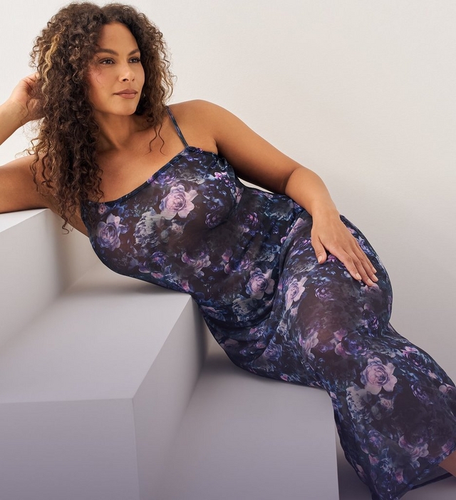 Close-up view of a woman with long curly brown hair reclining against a white angular platform, one hand resting near her head and the other resting on her hip. She wears a sheer navy slip dress with an allover purple and blue floral rose print and thin spaghetti straps. The background is a plain light gray wall.