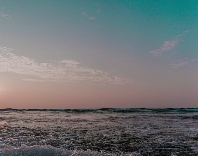 Photo of a beach and ocean at dusk. The sky transitions from teal at the top to pink and orange near the horizon, with wispy clouds in the upper left. The dark ocean water shows a line of breaking waves with white foam near the horizon, and rippled, foamy water in the foreground. A small distant landform or rock is faintly visible on the left horizon. No people or text appear in the image.